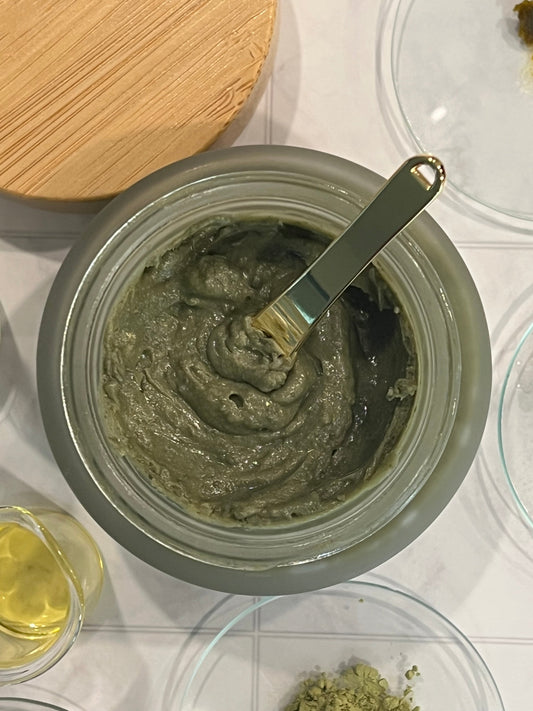 Making Oyabloom Matcha Clay Cleansing Balm — open jar of green balm with gold spatula on the formulation bench, matcha powder and oil alongside